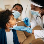 African American doctor with face mask examining boy's throat during a home visit.